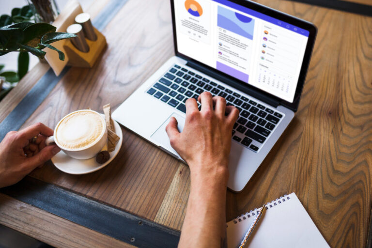 Pessoa em uma mesa usando notebook, com bloco de notas e uma xícara de bebida quente.